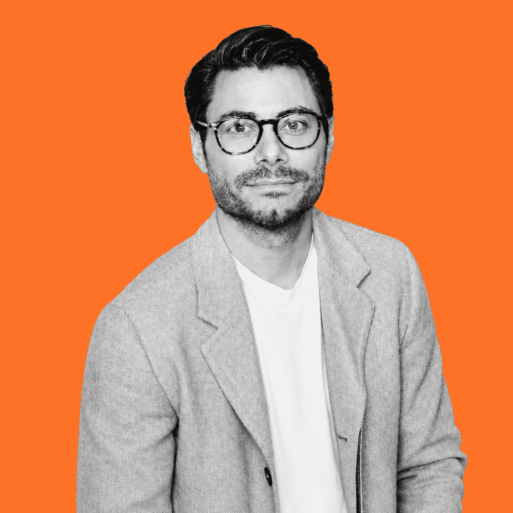 Black-and-white portrait of a man wearing glasses and a light blazer against a bright orange background.