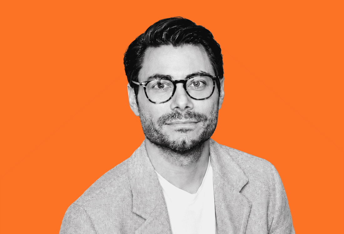 Black-and-white portrait of a man wearing glasses and a light blazer against a bright orange background.